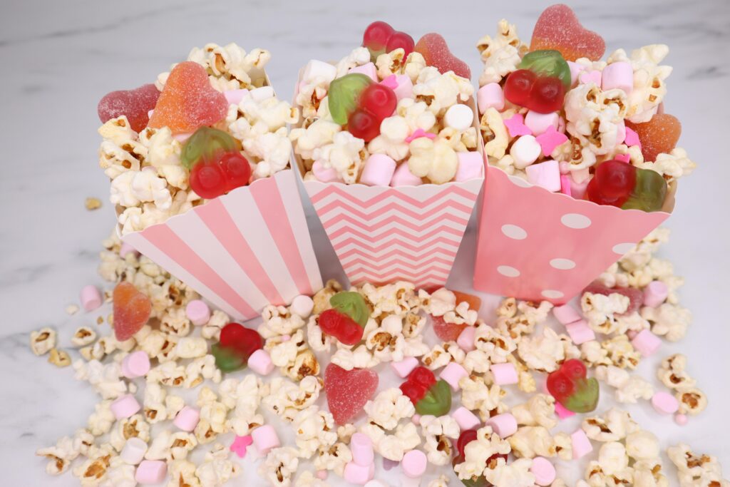 pexels-photo-18292635-18292635 Cheerful arrangement of popcorn, gummy candies, and marshmallows in decorative pink boxes on a white background.